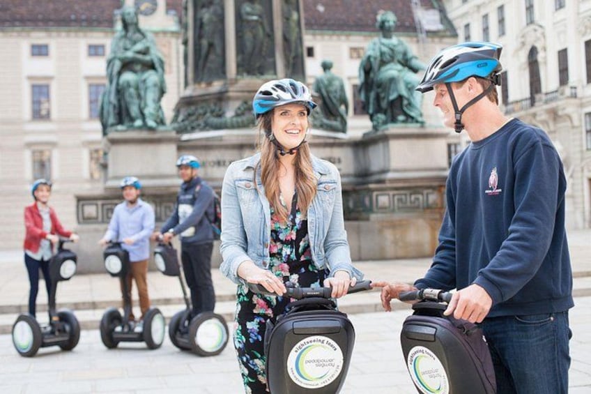 Een groep mensen op segways in een stedelijke omgeving met klassieke architectuur en beelden op de achtergrond. 3