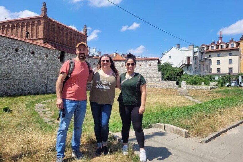 Three people standing in a grassy area with a stone wall and buildings in the background. 1