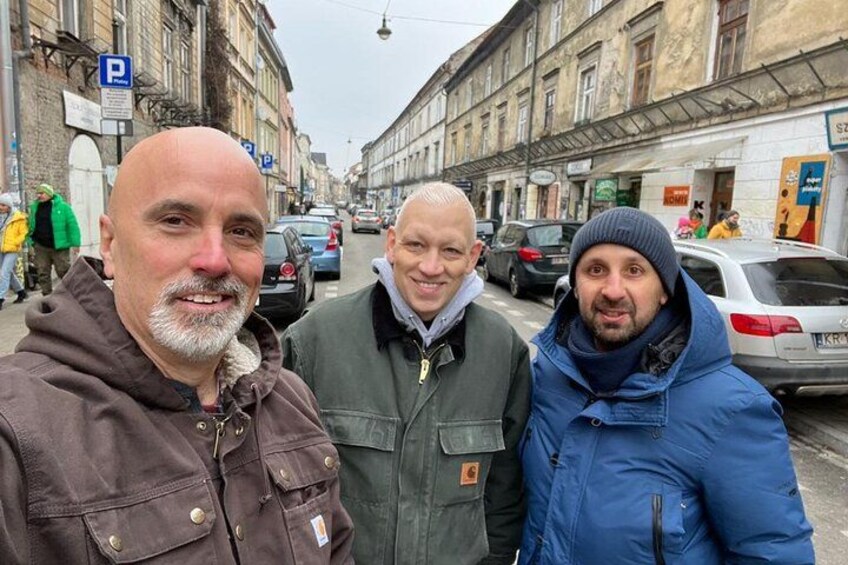Three men standing on a street with buildings and parked cars in the background. 2