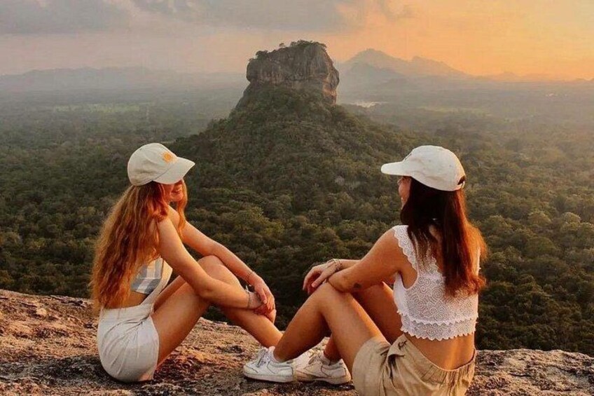 Two people sitting on a cliff overlooking a forested landscape with a prominent rock formation. 1
