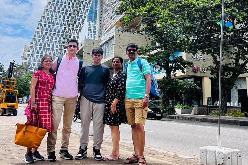 A group of people standing on a sidewalk with a modern building and a fountain in the background. 2