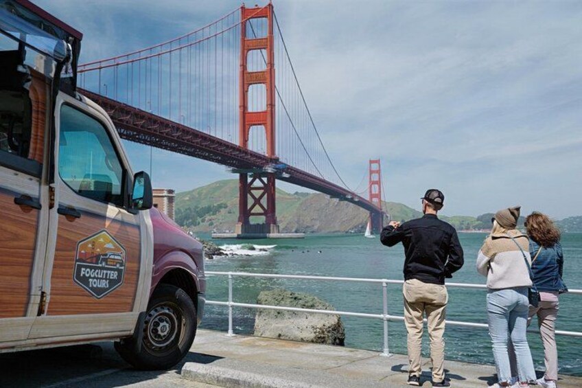 Un autobus en bois, portant l’inscription « Foghopper Tours » sur le côté, est garé au bord de l’eau, avec le pont du Golden Gate en arrière-plan. 1