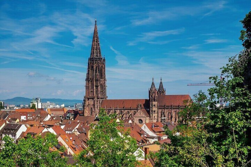 Une cathédrale dotée d’une haute flèche et d’une tour plus petite, entourée de bâtiments aux toits rouges et de verdure. 1