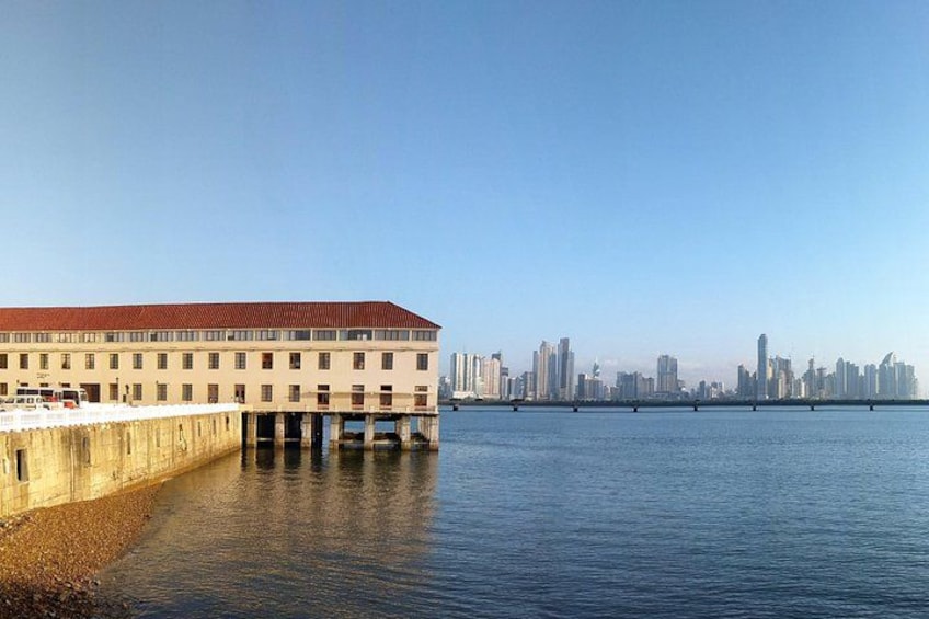 A waterfront building with a red roof, a pier extending into the water, and a city skyline in the background. 5