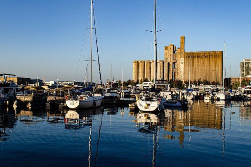A marina with several sailboats docked, a large industrial building, and calm water reflecting the boats. 2