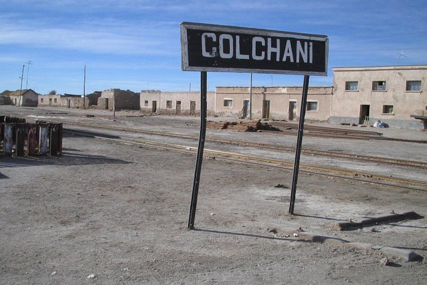 Señal que indica 'COLCHANI' en una zona rural con edificios y vías de tren. 2