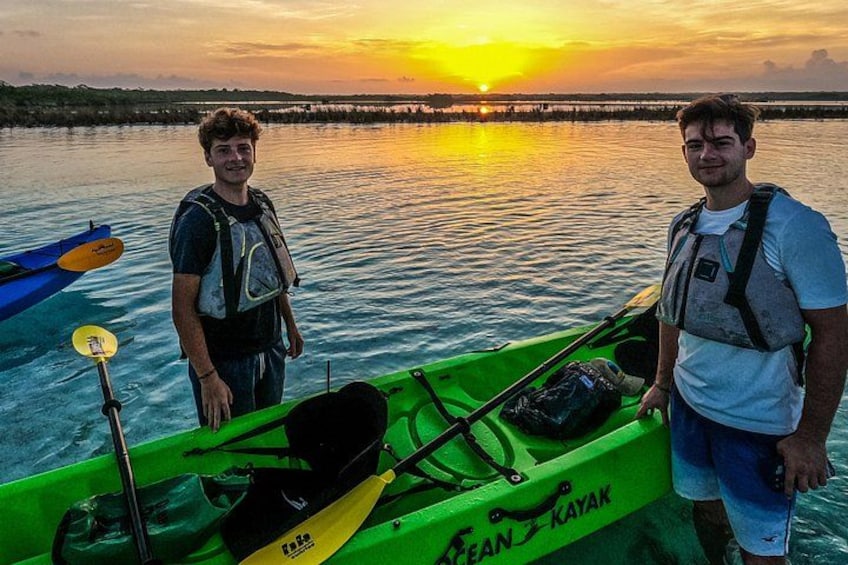 Due persone in piedi accanto a un kayak verde, con un tramonto sullo sfondo. 4