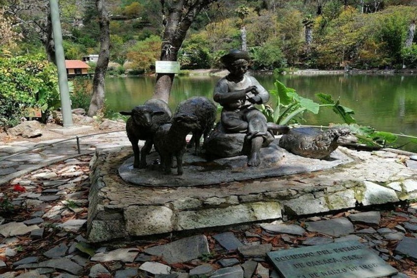 A statue of a man with two animals, a dog and a cat, in a park setting with a pond and trees in the background. 5