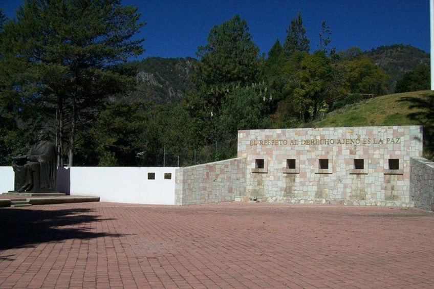 A stone wall with inscriptions, a statue, and a paved area with trees and hills in the background. 2