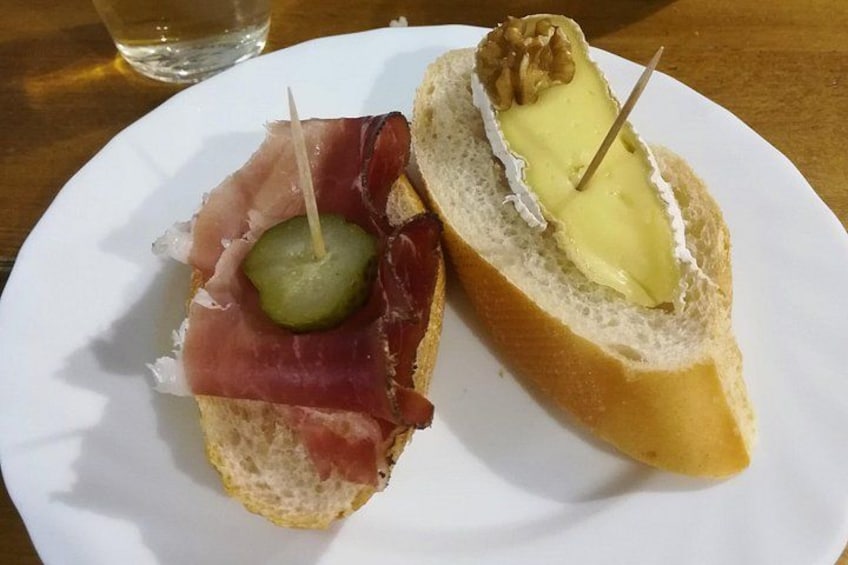 Deux sandwichs ouverts présentés sur une assiette blanche, l’un garni de jambon de Parme et d’un cornichon, l’autre de fromage et de noix. 2