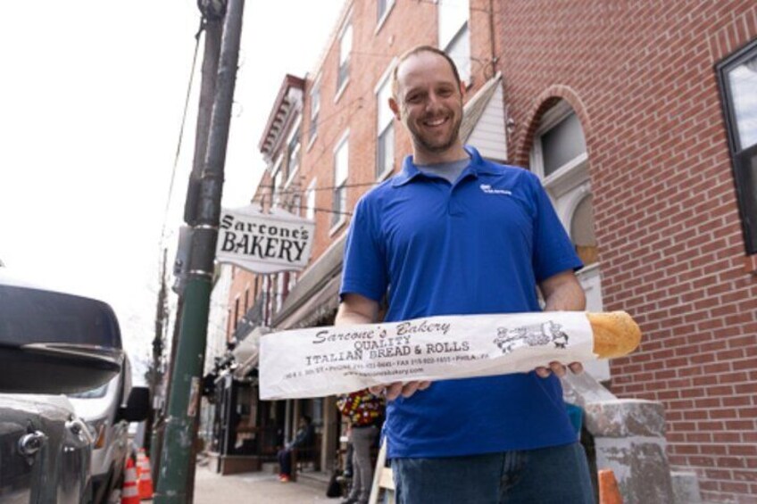 A man holding a large bread roll in front of a bakery sign. 2