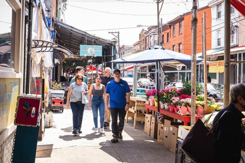 Un mercado callejero con gente caminando, puestos que venden flores y automóviles estacionados. 5