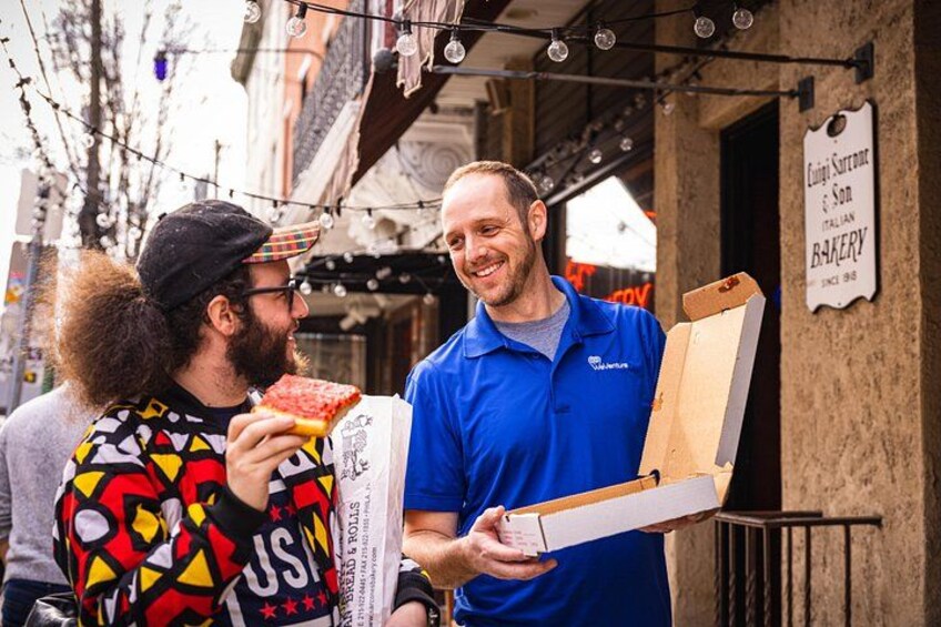 Two people, one holding a pizza box and the other holding a pizza slice, standing in front of a bakery with a sign that reads 'King Sorrento's Son Italian Bakery.' 2