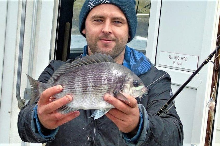 A person holding a fish, wearing a beanie and a jacket, with a fishing rod in the background. 1