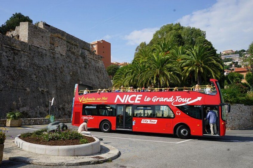 Ein roter, zweistöckiger Bus mit der Aufschrift 'NICE Le Grand Tour', der an einer Straße mit einer Steinmauer und Palmen im Hintergrund geparkt ist. 2
