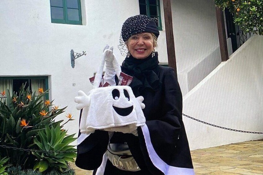 A person holding a ghost costume in front of a building with a green window and a plant with orange flowers. 3