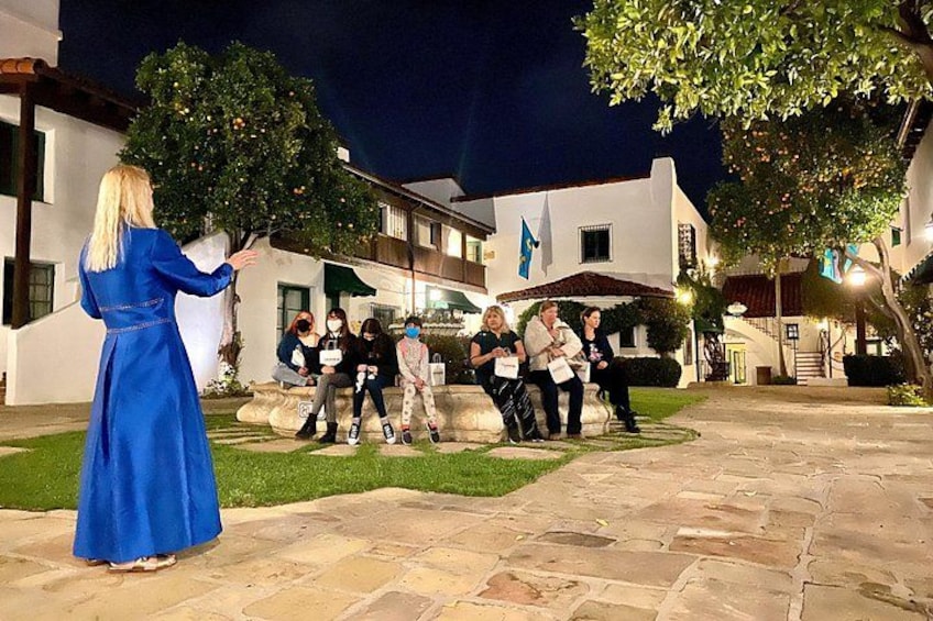 A woman in a blue dress standing on a stone patio, a group of people sitting on a bench, and a building with a white facade and blue flags. 2