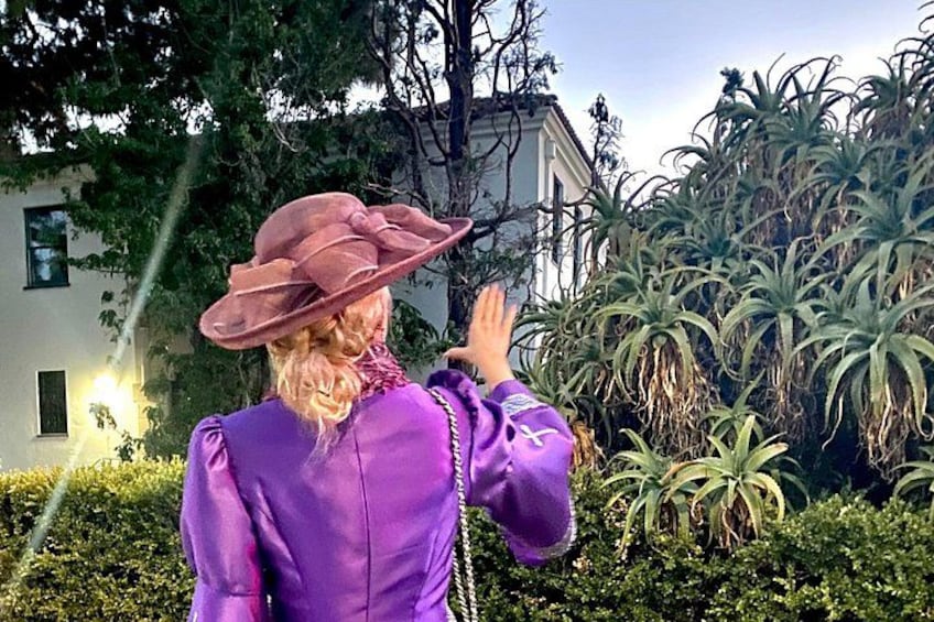 Une personne portant une veste et un chapeau violets, debout devant un bâtiment avec un jardin. 2