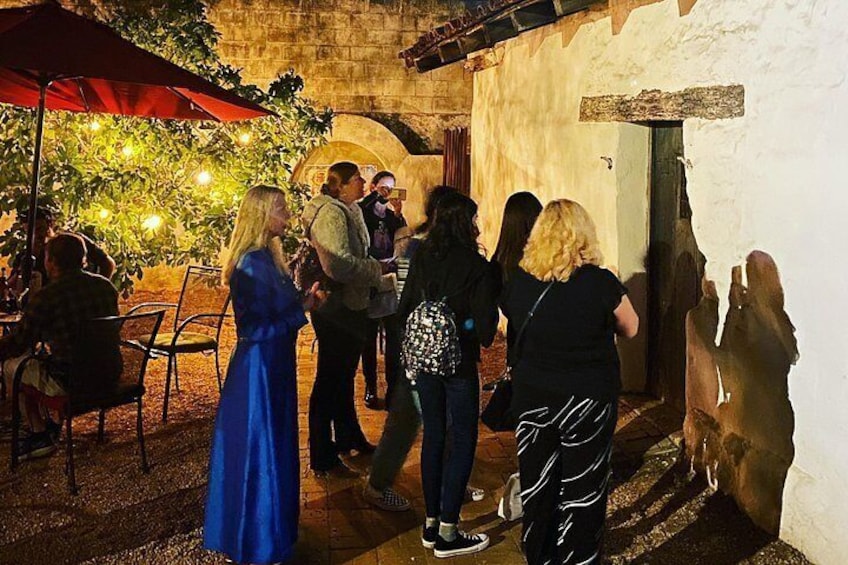 People gathered outside a building with a stone wall, under a red umbrella, and a tree with lights. 4