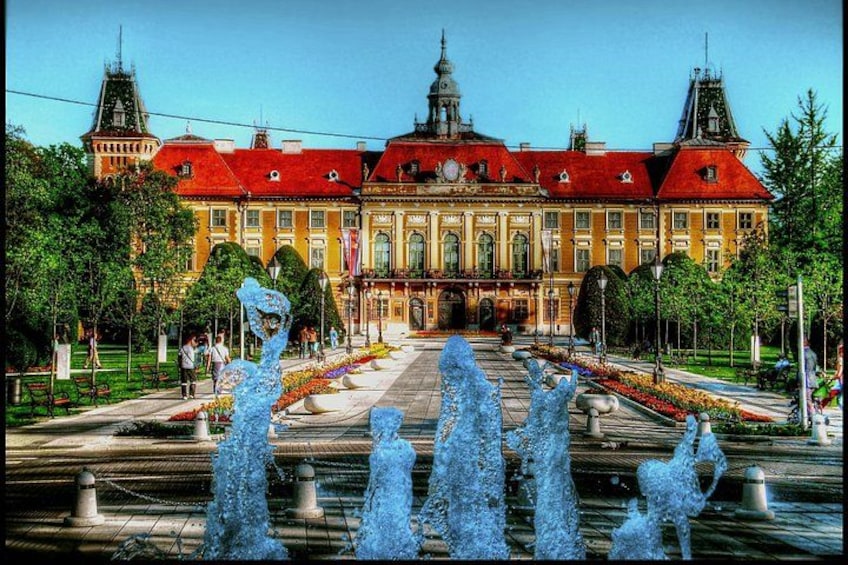A grand building with a red roof, a central dome, and a symmetrical facade. In front, there is a fountain with multiple water jets. 2