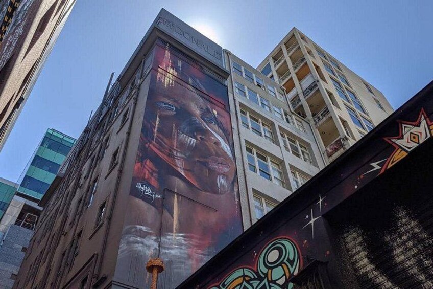Un edificio alto con un gran mural en la fachada, cielo despejado y una calle con grafitis. 3