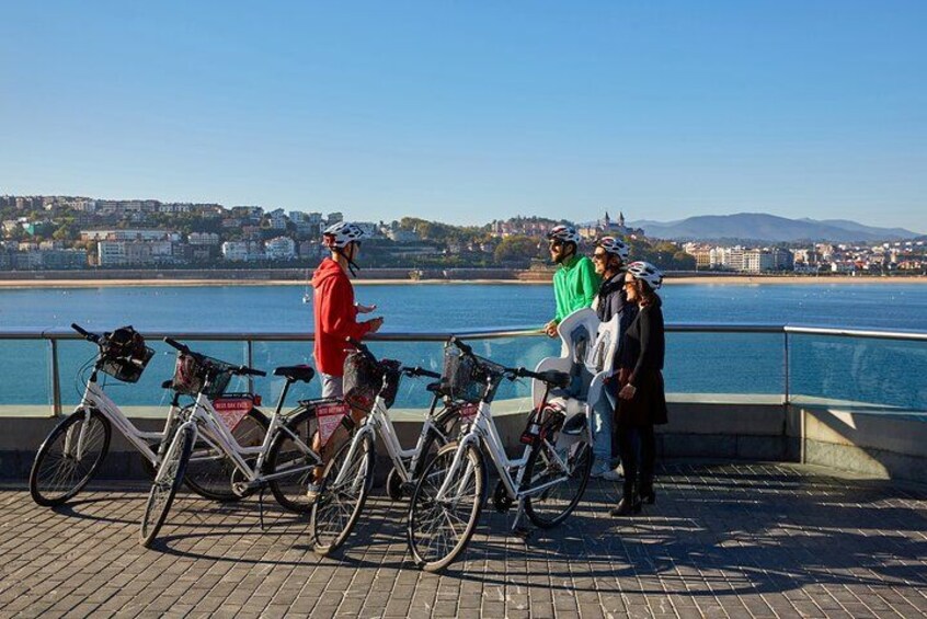 Un gruppo di persone in piedi accanto a biciclette lungo una passeggiata, con una città sullo sfondo. 2