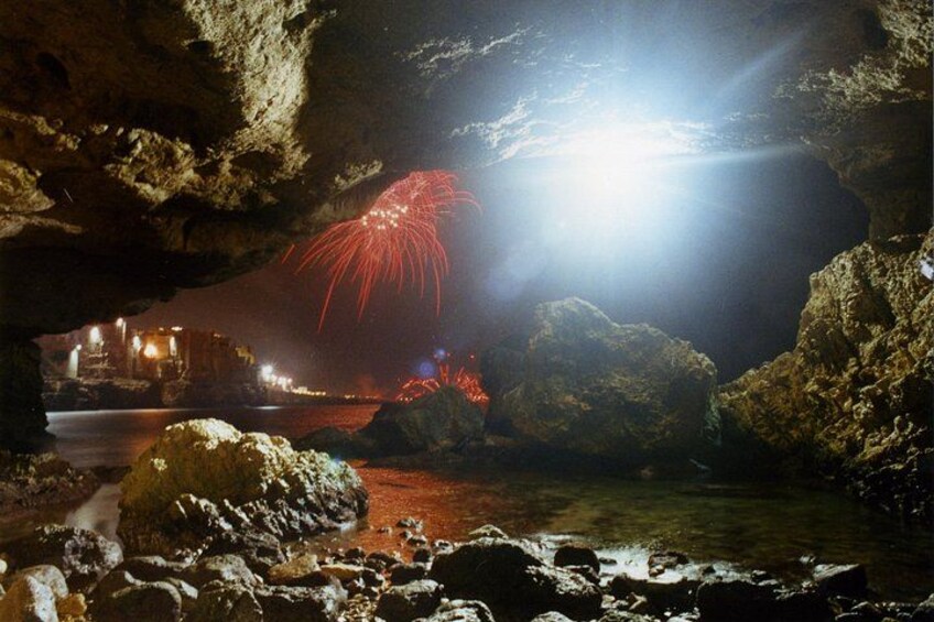 A rocky shoreline with a cave entrance, a body of water, and fireworks in the distance. 3