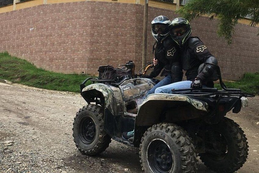 Two people on an ATV with a brick wall and greenery in the background. 4