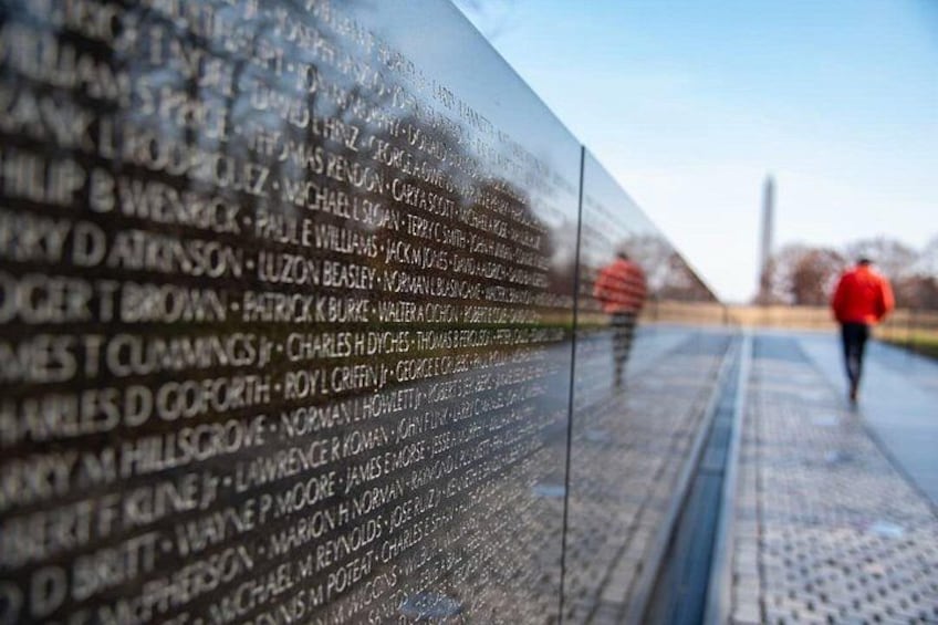 A memorial wall with names inscribed, a person in red walking in the background, and a clear sky. 3