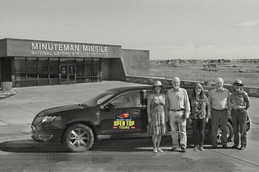A black and white photo of a group of people standing in front of the Minuteman Missile National Historic Site Visitor Center with a car displaying the Black Star Tours logo. 4