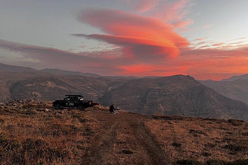 A jeep on a dirt path with a person walking away, surrounded by mountains and a sunset sky. 3
