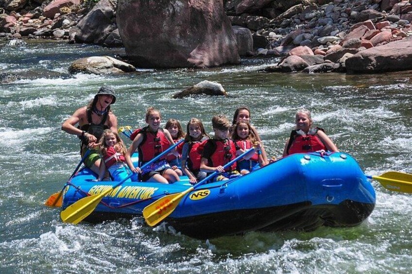 A group of people rafting on a blue raft with yellow paddles, surrounded by a river and rocks. 2