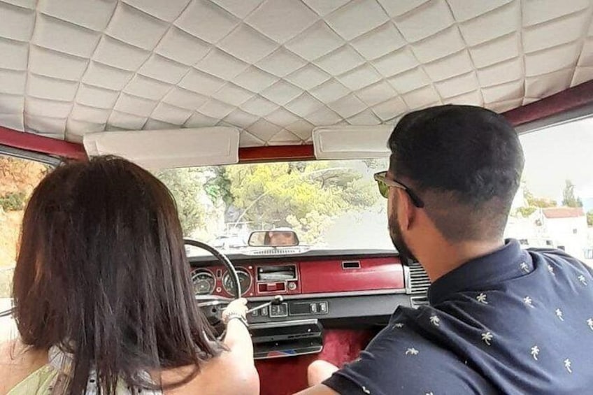 Het interieur van een oldtimer met een vrouw achter het stuur en een man op de passagiersstoel. 1