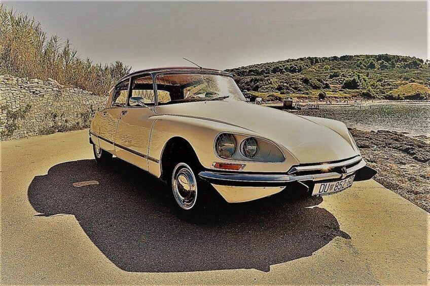 Une voiture ancienne est garée sur une route côtière, avec un mur de pierres et une plage en arrière-plan. 1