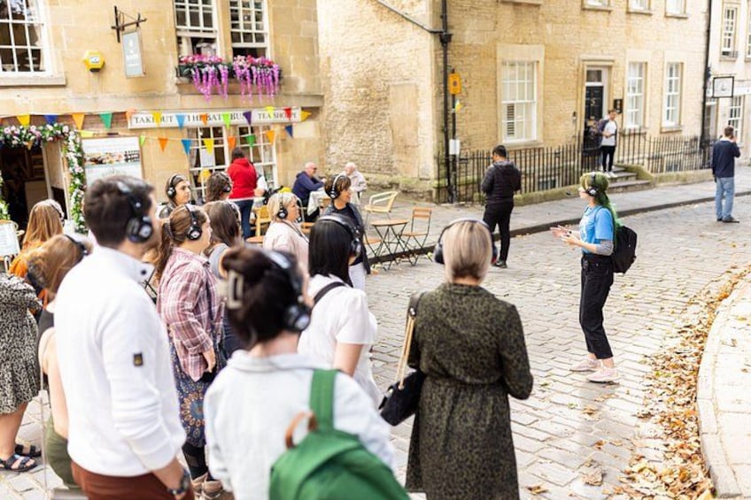 A group of people gathered on a cobblestone street, some wearing headphones, in front of a building with a sign that reads 'TAKI OUT THEATRE.' 1