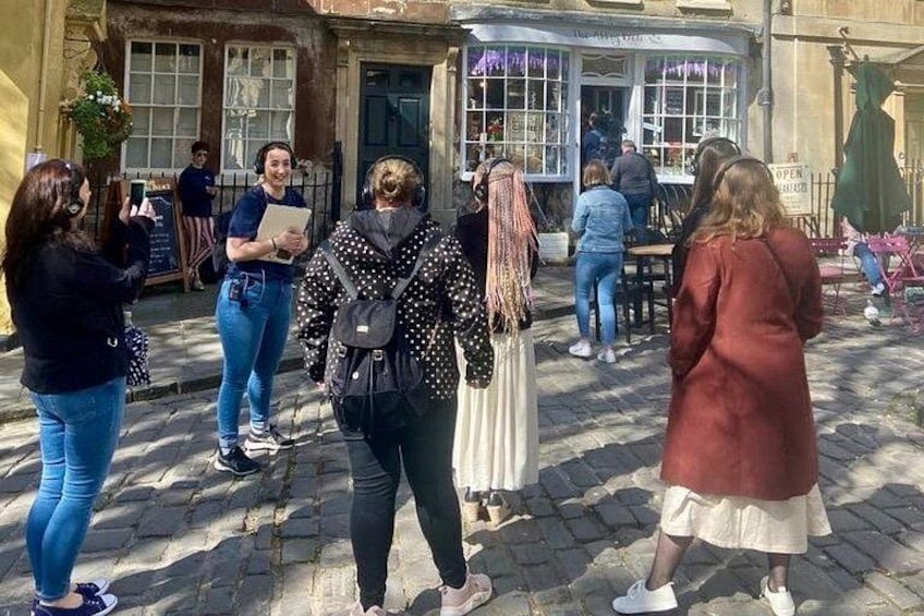 A group of people standing on a cobblestone street, some holding papers, with a building and a shop in the background. 5