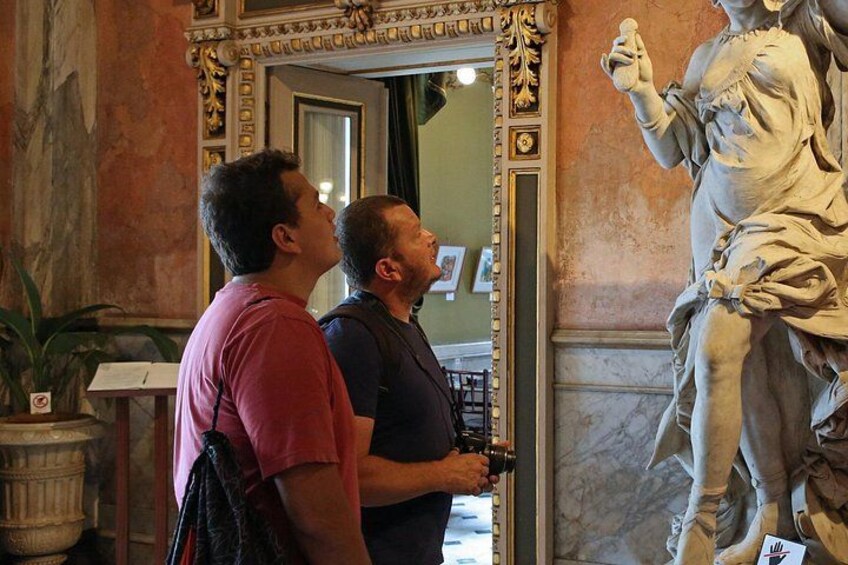 Two people observing a marble statue in a room with ornate decorations. 1