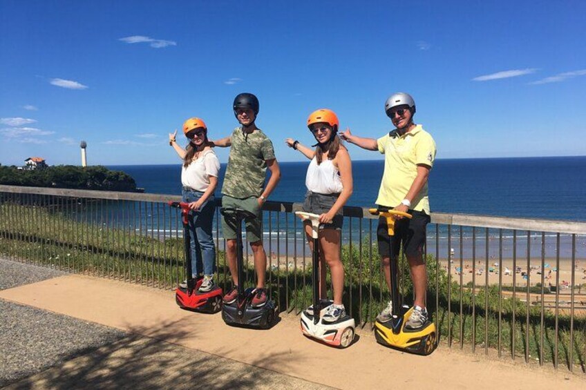 Vier personen op hoverboards langs een kustweg, met een strand en oceaan op de achtergrond. 2