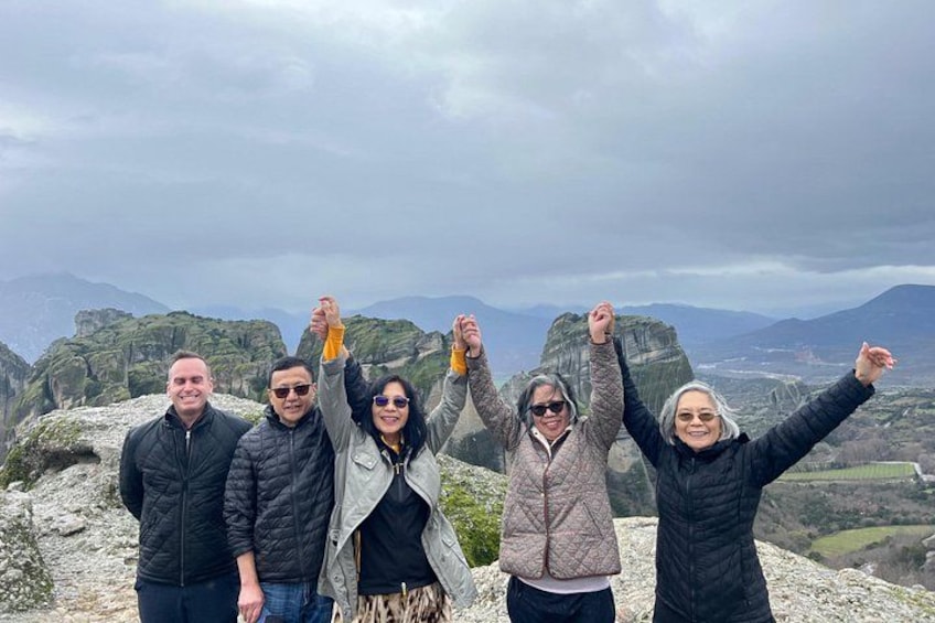 Un grupo de personas de pie sobre un saliente rocoso, con los brazos levantados, contemplando un paisaje montañoso. 2