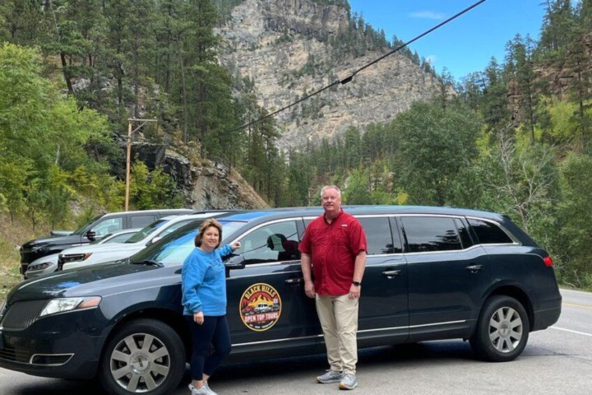 A black SUV with a logo on the side parked on a road, surrounded by trees and a rocky mountain in the background. 1