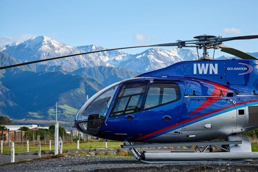 Un hélicoptère bleu et rouge, portant les inscriptions « IWN » et « GCH Aviation » sur le côté, est stationné avec une chaîne de montagnes en arrière-plan. 2