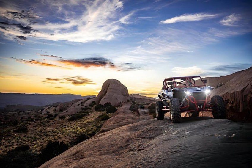 A red off-road vehicle on a rocky terrain with a large rock formation in the background and a sunset sky. 4