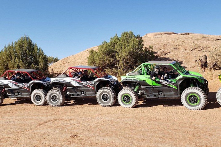 Four off-road vehicles parked on a dirt surface with a rocky hill and trees in the background. 1