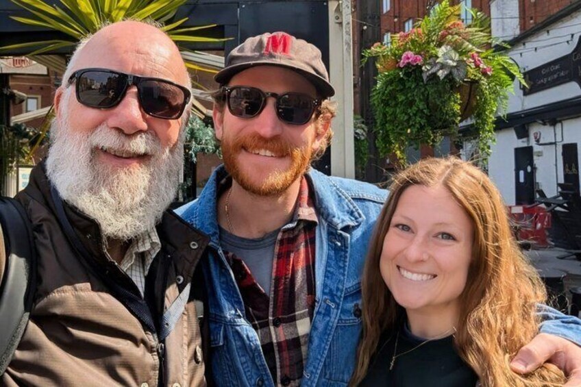 Three people standing outdoors with buildings and a hanging plant in the background. 3