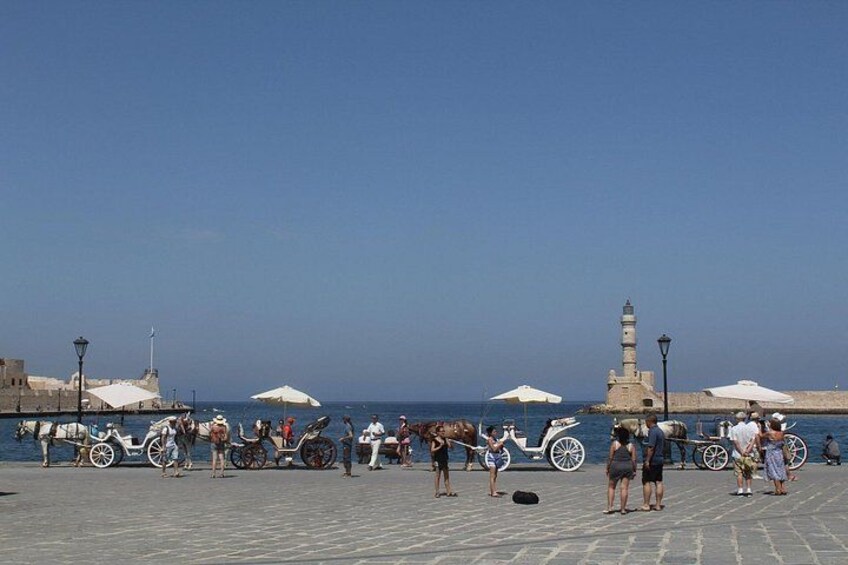 A seaside promenade with horse-drawn carriages, a lighthouse, and people walking. 2