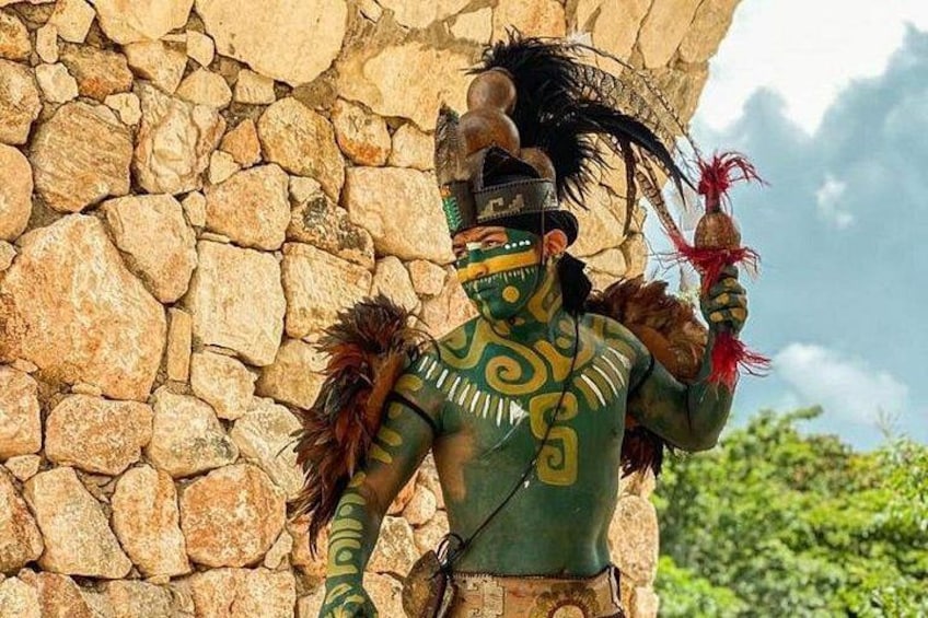 A person in traditional attire with a headdress and feathered accessories, standing in front of a stone wall. 3
