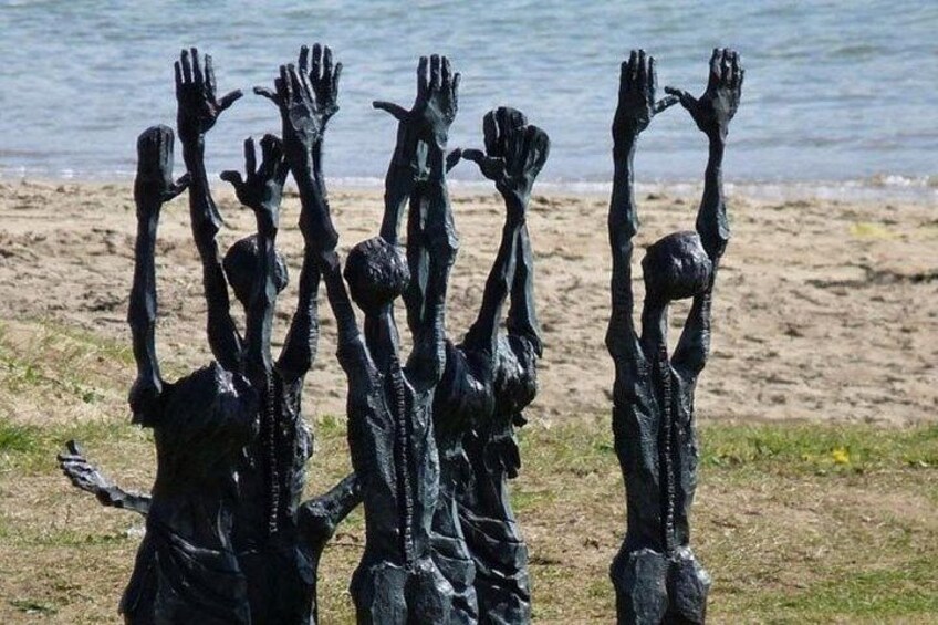 Un ensemble de sculptures en bronze représentant des personnages les bras levés, situé près d’une plage. 1