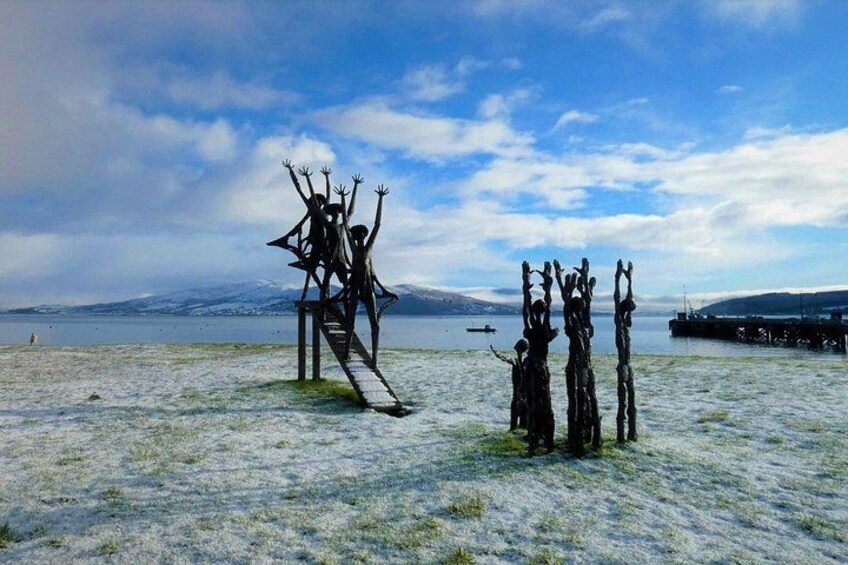 Escultura de figuras con brazos alzados sobre una plataforma, masa de agua y una cadena montañosa distante. 3