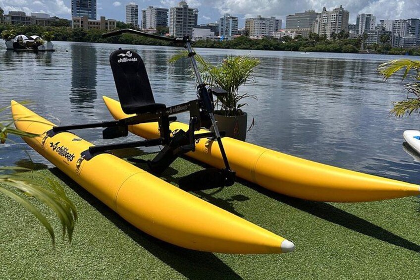 Un kayak giallo con un sedile nero e una pianta sulla riva di un lago, con sullo sfondo la skyline di una città. 4