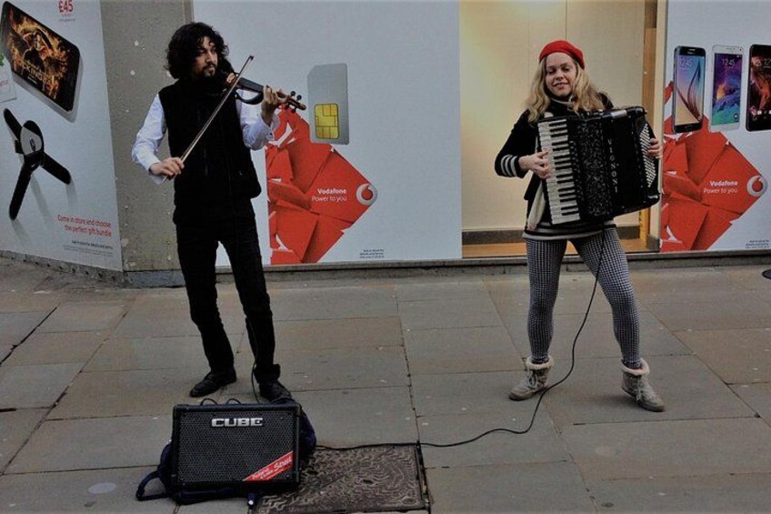 Two people playing musical instruments, a violin and an accordion, on a sidewalk with a speaker and a poster in the background. 3